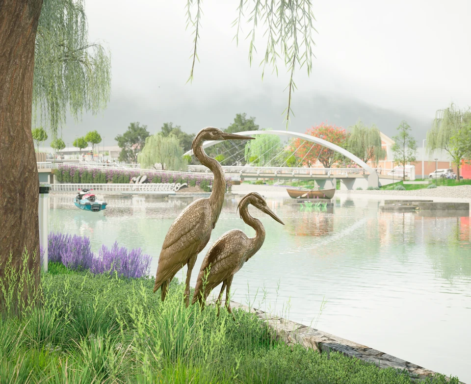 Dos esculturas de garzas entre pastos a orillas de la laguna; al fondo, un puente de arco y botes.